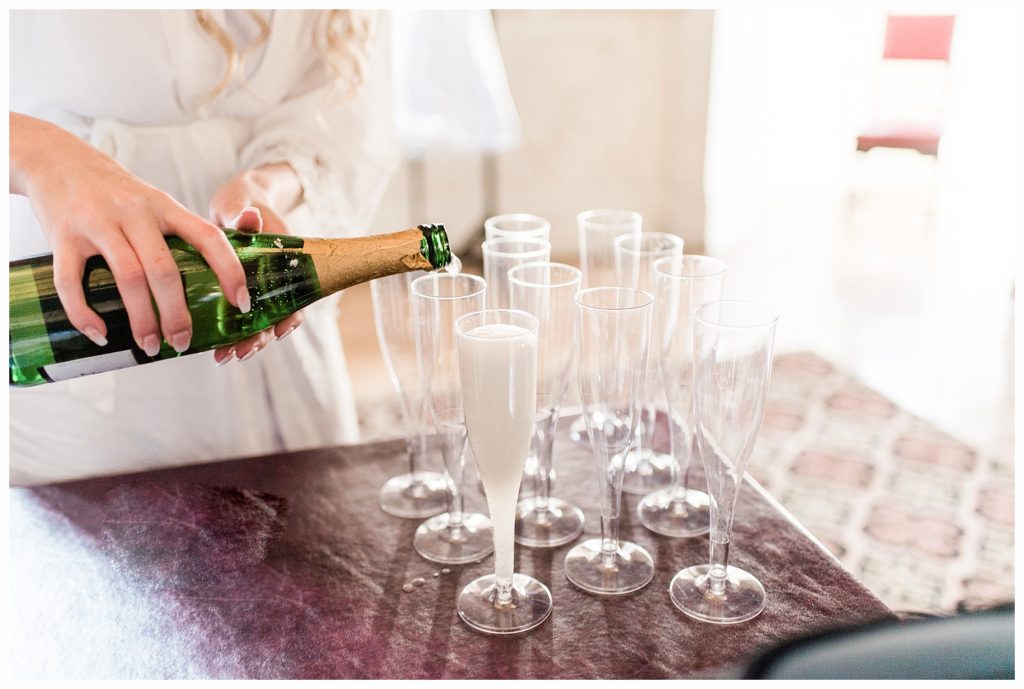Colored image on a bride from the chest down pouring champagne into flutes for her bridesmaids. She's wearing a white robe. Photo by Kayla Lee.