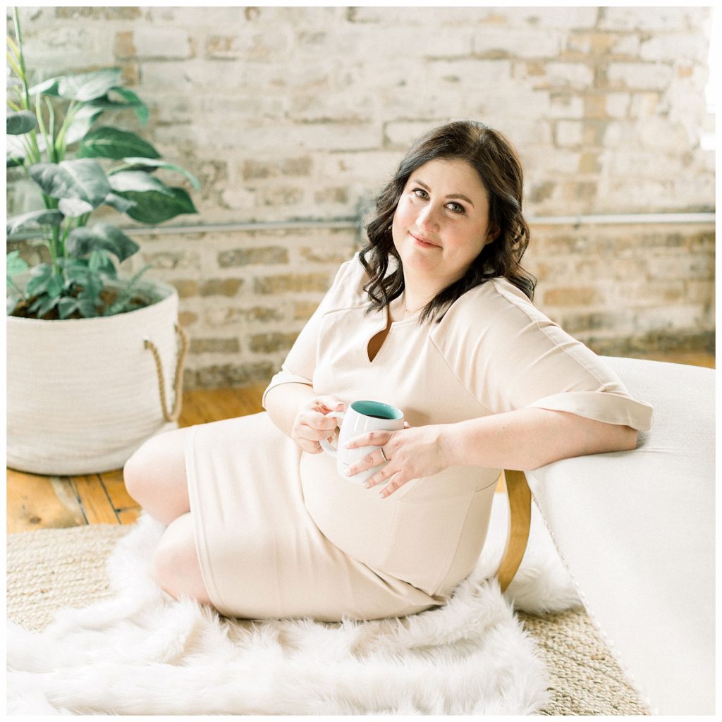 Female in nude dress sits on a fuzzy rug drinking a cup of coffee on National Coffee Day.