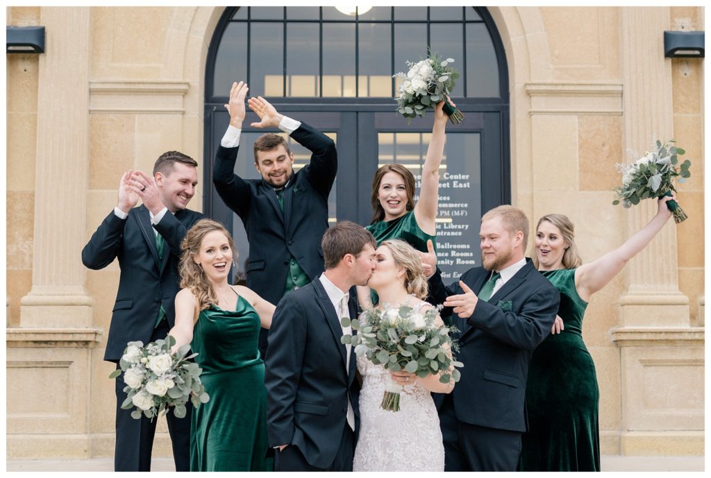 Wedding party wearing black suits and green velvet dresses cheer on the couple as they kiss. This post is about the best wedding party gifts. Photo by Kayla Lee.
